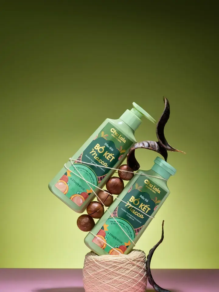 Chai dầu gội và dầu xả Chu Lala Bồ Kết Macca nằm nghiêng chồng lên nhau, cạnh quả bồ kết và hạt mắc ca trên nền xanh olive