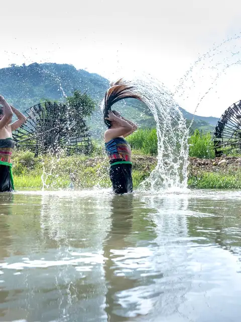 Ba cô gái đứng giữa dòng suối, cùng hất mái tóc dài ướt tạo vòng nước, sau lưng là núi xanh.