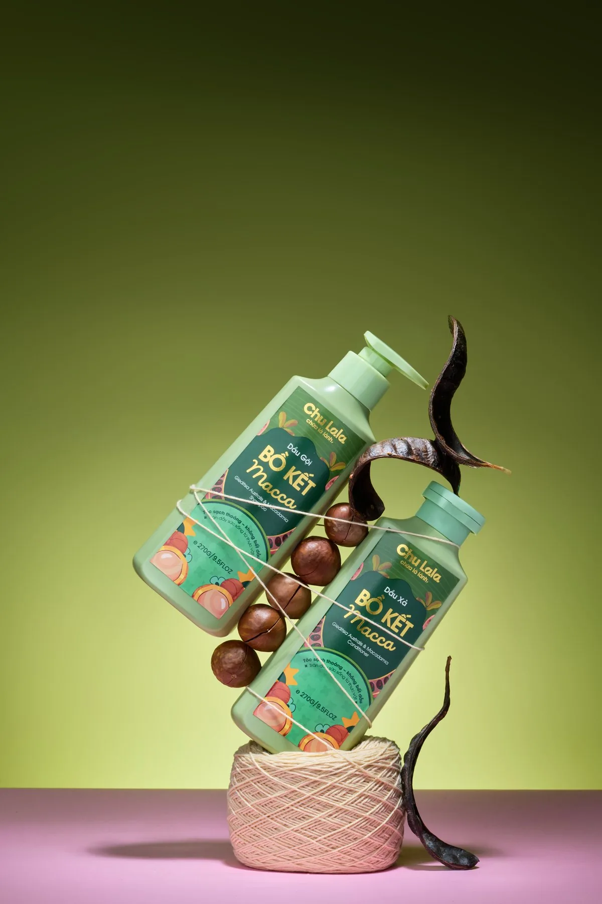 Chai dầu gội và dầu xả Chu Lala Bồ Kết Macca nằm nghiêng chồng lên nhau, cạnh quả bồ kết và hạt mắc ca trên nền xanh olive