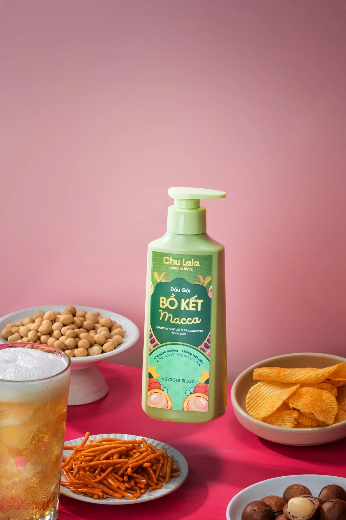 Chai dầu gội Chu Lala Bồ Kết Macca trên nền hồng, cạnh vài món ăn vặt